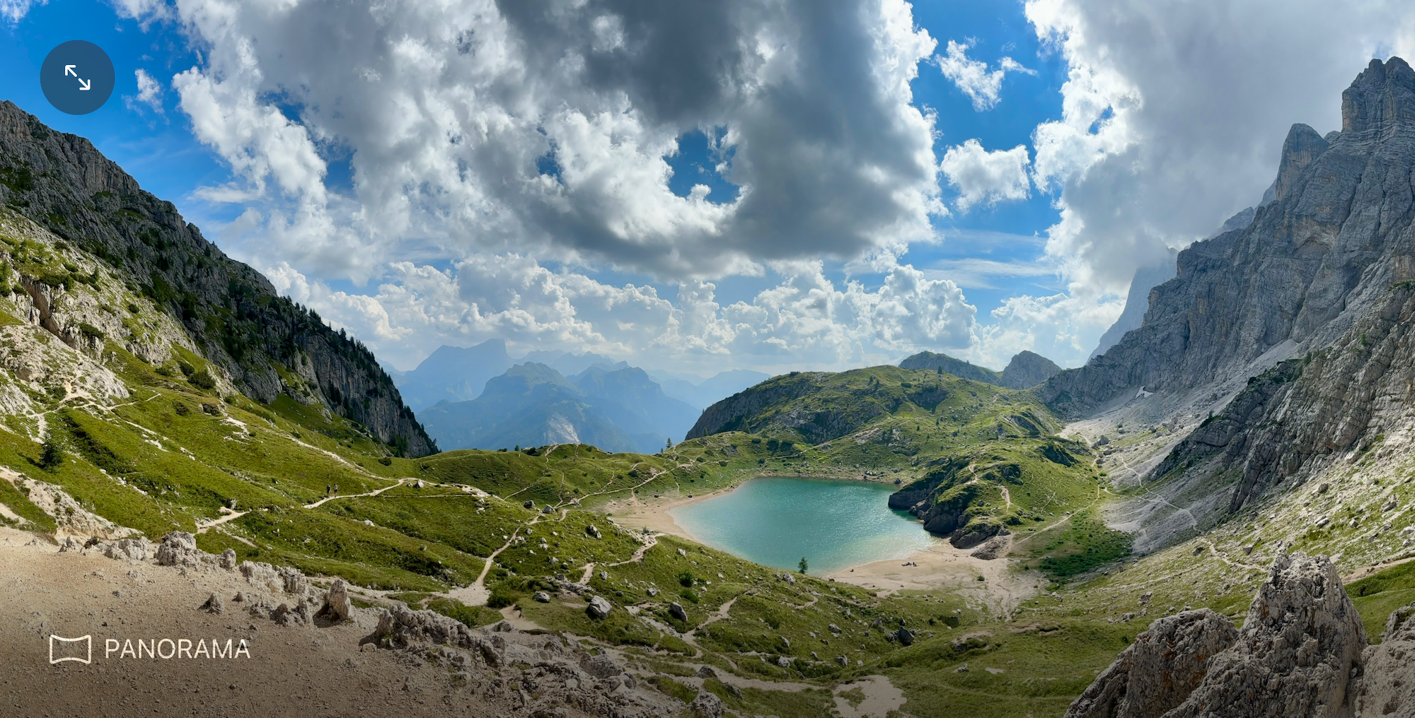 Photo of valley surrounded by mountains, an open cloudy sky, and a lake towards the right center.